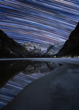 View of star trails over the frozen Durrensee lake with snow-covered mountains in the background at night in Toblach, Trentino-Alto Adige, Italy.