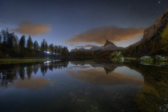 View of Lago Federa at night with a starry sky reflecting in the calm water, surrounded by autumn larch trees and mountain peaks in Cortina d'Ampezzo, Veneto, Italy.