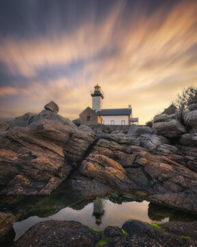 View of Phare de Pontusval lighthouse at sunset with dramatic long-exposure clouds and rugged granite rocks in the foreground in Plouneour-Brignogan-plages, Bretagne, France.