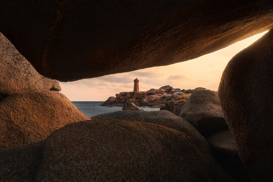 View of Phare de Men Ruz framed by large granite boulders on the coast at sunset in Perros-Guirec, Brittany, France.