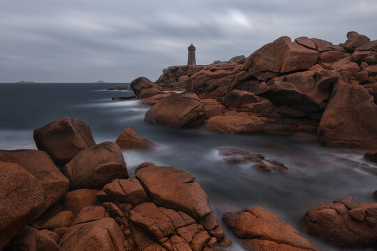 View of the Phare de Men Ruz lighthouse standing on the rugged pink granite rocks of the coast with long exposure sea waves in Perros-Guirec, Brittany, France.