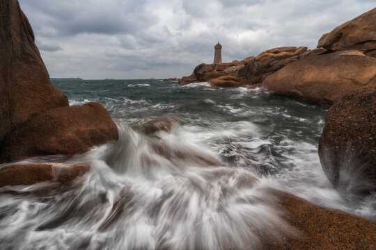 View of Phare de Men Ruz lighthouse standing on pink granite rocks with waves crashing against the shore under a cloudy sky in Perros-Guirec, Brittany, France.