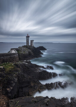 View of Le Phare du Petit Minou lighthouse standing on a rocky cliff with a stone walkway and long exposure waves crashing against the shore in Plouzane, France.