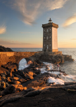 View of Kermorvan Lighthouse on a rocky coastline with crashing waves and a person fishing at sunset in Le Conquet, Bretagne, France.