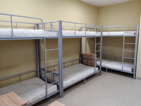 Bunk beds arranged in a dormitory room with beige walls and gray flooring, featuring empty mattresses and wooden furniture for storage
