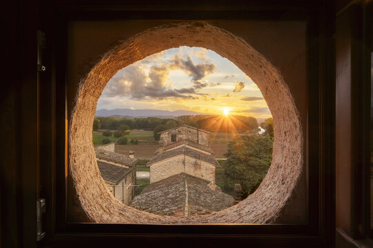 View of the village of San Giacomo at sunset framed by a rustic circular stone window, featuring historic stone rooftops and golden hour light in San Giacomo, Umbria, Italy.