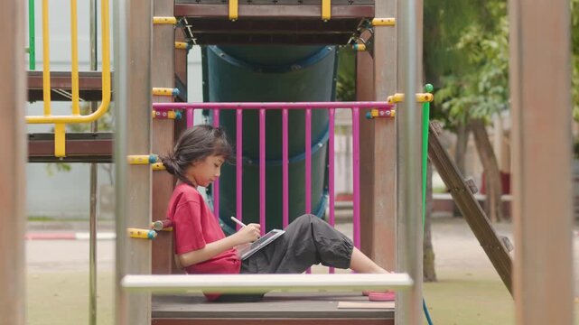 Asian kid enjoying a relaxing hobby, drawing digital art on tablet at outdoor school playground. Selective focus highlights creative elementary student spending personal leisure time with technology.