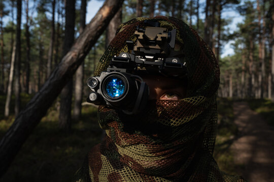 Female soldier in camouflage scarf looking through night vision monocular with glowing blue lens during moonlight night operation in forest