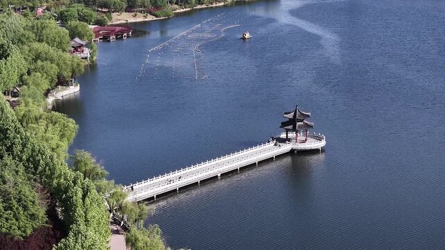 Aerial view of a traditional Chinese pavilion on a long lakeside walkway with fishing nets spread across the calm water surface