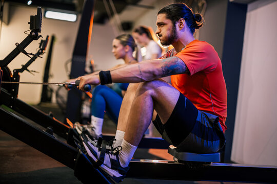 Side view of two people training on indoor rowing ergometers during a group fitness class in a modern gym.