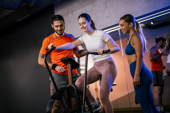 A male coach stands in front of a group of people on rowing machines, cheering them on and directing a high-intensity group class.
