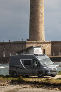 View of a modern grey camper van with a pop-top roof parked near the historic Gatteville-le-Phare lighthouse on the rocky coast of Gatteville-le-Phare, Normandy, France.