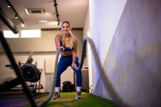 Full body shot of a beautiful athletic woman performing a high-intensity battle ropes routine on green artificial grass inside a contemporary fitness center.