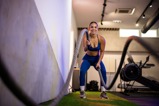 Side angle shot of an athletic woman in a deep squat position working out with battle ropes, targeting core stability and lower body endurance.