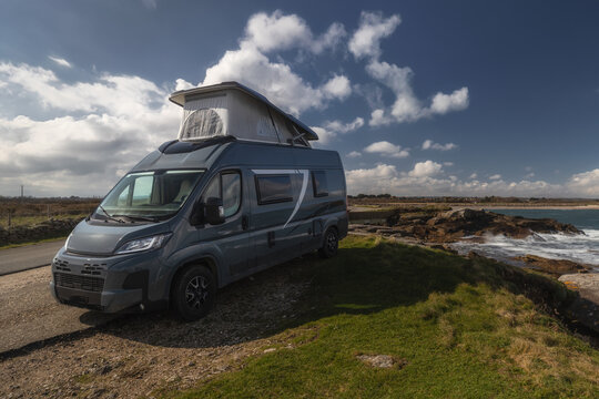 View of a grey camper van with a pop-up roof parked on a coastal road near the rocky shoreline and sea in Gatteville-le-Phare, Normandy, France.