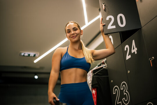 Medium shot of a beautiful fit woman looking directly into the camera with a confident smile, standing in a contemporary gym locker room with industrial design.