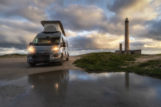 View of a camper van parked near the historic Gatteville-le-Phare lighthouse at sunset, with a reflection in a puddle on the ground in Gatteville-le-Phare, Normandy, France.