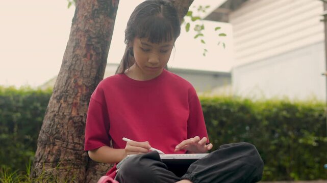 Selective focus captures the face of an Asian kid relaxing under a tree. With clear copy space, she enjoys drawing digital art and studying on a tablet during her casual outdoor hobby.