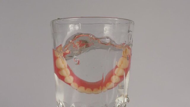 A removable full denture falls into a glass of clean water causing a splash. Demonstration of daily hygienic care and cleaning process for false teeth at home