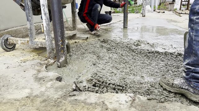 Male hands in protective gloves uses trowel to spread and level fresh cement in yard. Another worker distributes and moves the mixture with a shovel preparing the material during construction process