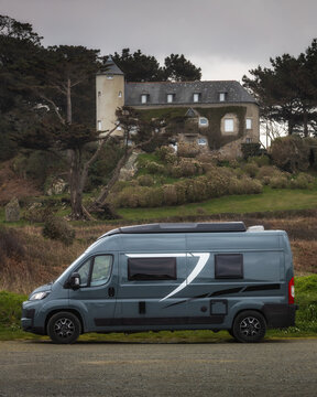 View of a modern grey camper van parked in front of a historic stone house with a turret, surrounded by trees and greenery in Normandy, France.