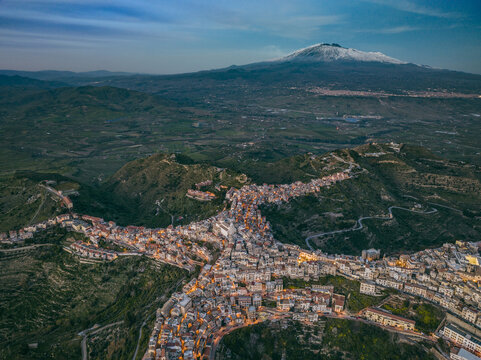 Aerial view of the town of Centuripe, featuring its unique human-like shape illuminated at dusk with Mount Etna in the background, Centuripe, Sicily, Italy.