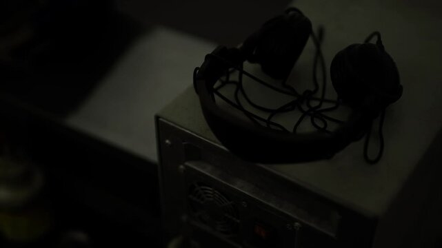 Headset resting on computer chassis with coiled cable, metal case surface and blurred equipment background, dim studio lighting evokes monitoring or salvage