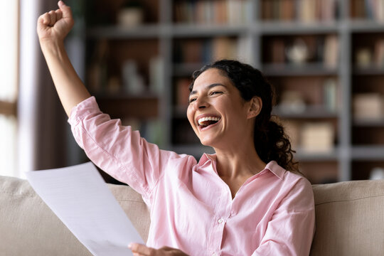 Smiling woman raises her fist while holding paperwork on a sofa at home. Good news, personal success, happy achievement.