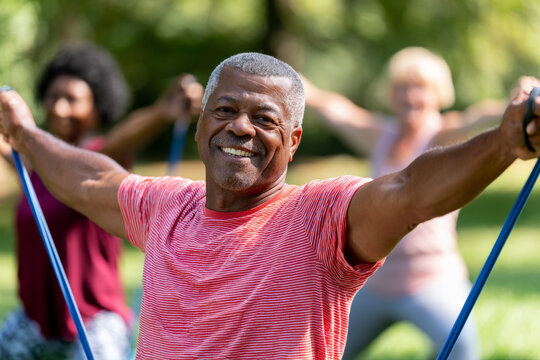 Senior Black man raises a resistance band during an outdoor group workout. Active aging, strength training, park fitness.