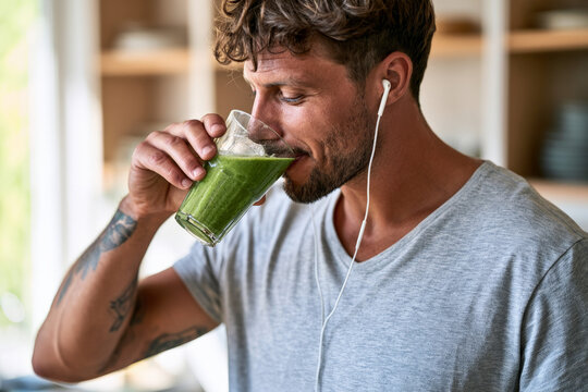 Young bearded man drinks a green smoothie while wearing earbuds at home. Healthy beverage, daily nutrition, casual wellness.