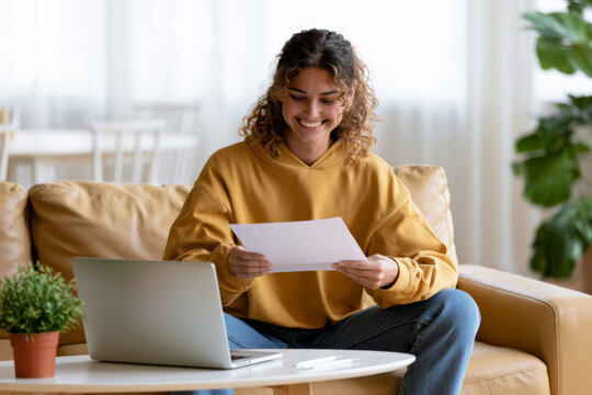 Young curly woman reads a letter beside a laptop while sitting on a sofa. Good news, personal success, positive result.