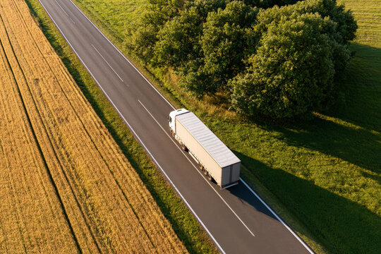 White freight truck travels along a rural road between fields and trees. Road transport, cargo delivery, regional logistics.