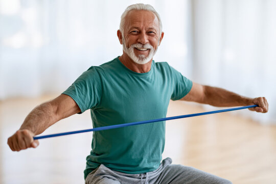 Smiling senior man exercises with a blue resistance band at home. Active aging, strength training, healthy mobility.