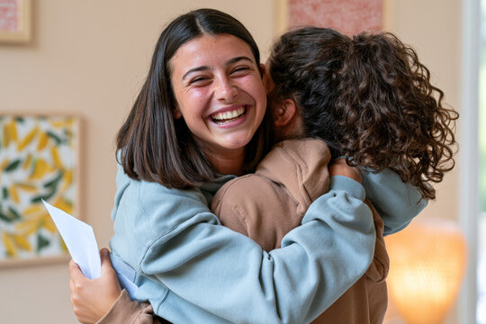 Teenage girls laugh during a warm embrace while holding papers at home. Friendship support, positive news, happy achievement.