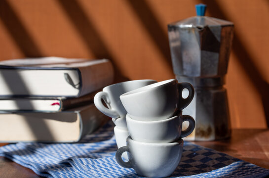 Espresso cups and an old coffee maker are sitting on a cloth in front of some books. The background is brown. The light is shining on the cups.