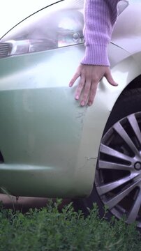Woman's hand examines scratches and cracks on a light green car bumper after an accident. Outdoor close-up showing wheel and bushes.