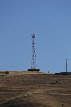 A red and white cell tower rises from a hedge-ringed hilltop into empty blue sky, rural mobile coverage in vertical format, Romania.