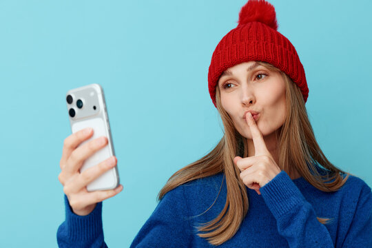 A woman takes a selfie in a cozy winter outfit, finger to lips in a hush gesture. A bright blue background emphasizes a private moment of secret self expression.