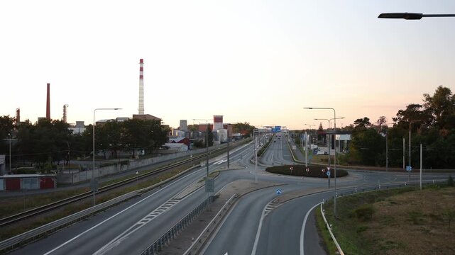 Timelapse shows sparse traffic moving along a wide urban road beside railway tracks and industrial buildings during sunset as passing cars create smooth light trails afar