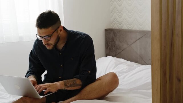 Focused man in casual clothes and glasses works on laptop while sitting on bed in bright modern bedroom. Horizontal 4k footage
