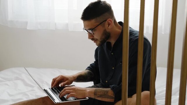Focused man in casual clothes and glasses works on laptop while sitting on bed in bright modern bedroom. Horizontal 4k footage