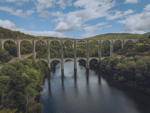 Aerial view of Cize-Bolozon viaduct spanning the Ain River, surrounded by lush green forests and rolling hills under a blue sky with scattered clouds in Corveissiat, Auvergne-Rhone-Alpes, France.