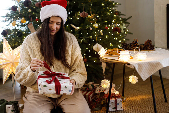 Happy Woman in Santa Hat Opening Christmas Gift Box Sitting Near Christmas Tree and Cozy Fireplace in festive living room. Happy holidays