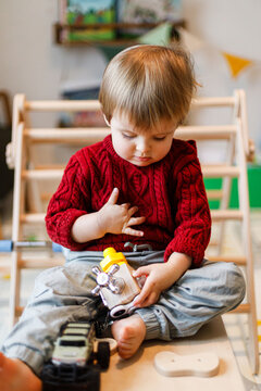 Cute toddler boy in red sweater sitting barefoot and playing with wooden sensory busy cube toy indoor in kid playroom