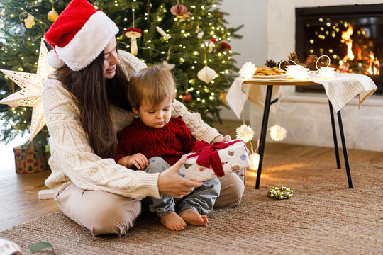 Mother in Santa Hat and Little Son Opening Christmas Present Together Near Fireplace and decorated xmas Tree in festive living room. Merry Christmas