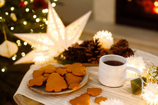 Plate of homemade gingerbread cookies and hot tea on table near illuminated star and Christmas tree. Cozy xmas atmosphere. Hygge winter