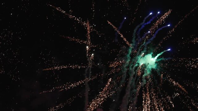 Spectacular fireworks explode across the night sky, sending bright colorful rockets and showers of glowing sparks upward through clouds of smoke. The dynamic display creates a festive atmosphere