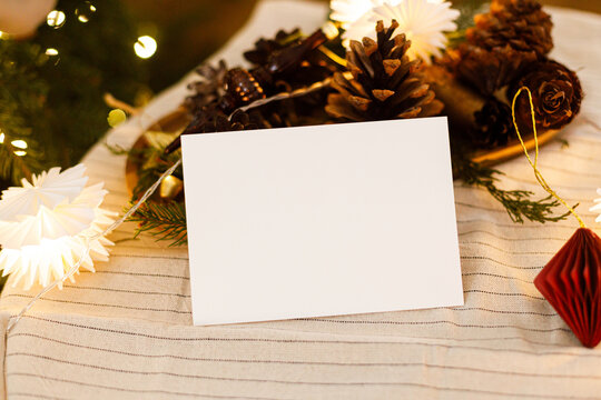 Festive blank paper card mockup template on decorated Christmas table with pine cones, ornaments and lights. Xmas card mock up with copy space