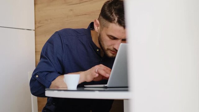 Focused man works on laptop at home kitchen table, concentrating on screen while drinking morning coffee. Digital nomad at home office. Horizontal 4k footage