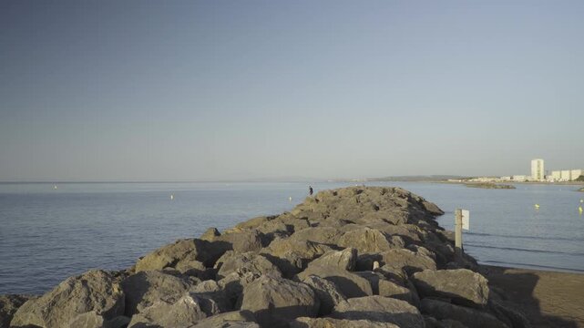 France, Valras Plage August 08, 2026 &mdash; Wide shot of a long stone breakwater pier extending into the calm blue Mediterranean sea with a city shoreline and buildings in the background under clear sky